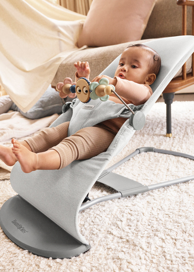 Baby in a gray baby bjorn bouncer bliss with a toy, sitting on a carpeted floor.